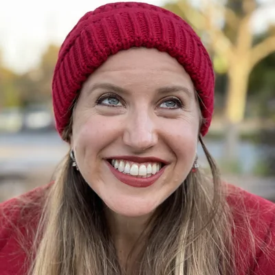 A close-up portrait of a smiling woman wearing a red knit beanie and a red coat, looking upward and to her right. She is outdoors with a softly blurred background of trees and a street.