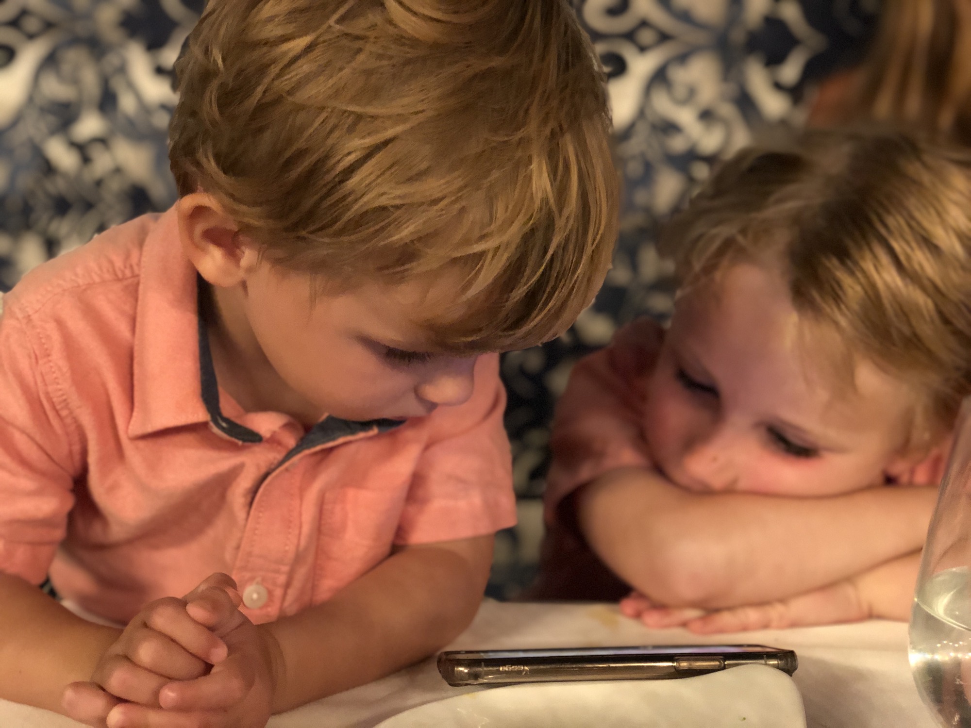 Two young children in matching pink shirts are sitting at a table indoors, looking down at a smartphone screen.