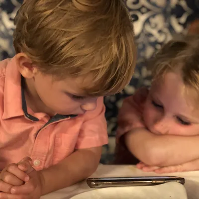 Two young children in matching pink shirts are sitting at a table indoors, looking down at a smartphone screen.