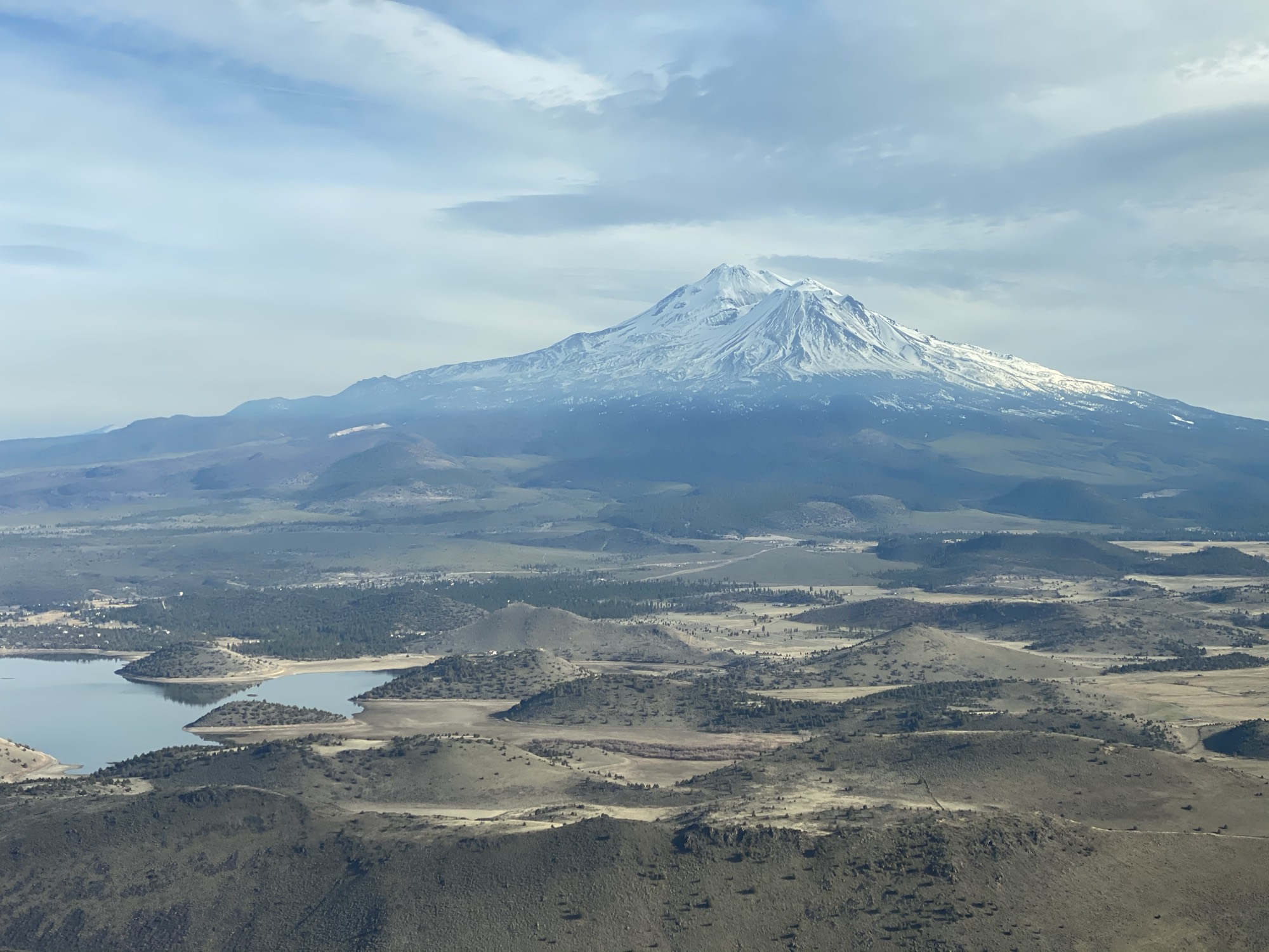An aerial view captures a large, snow-capped mountain rising above a vast landscape of rolling hills, fields, and a body of water under a partly cloudy sky.