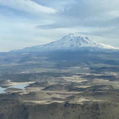 An aerial view captures a large, snow-capped mountain rising above a vast landscape of rolling hills, fields, and a body of water under a partly cloudy sky.