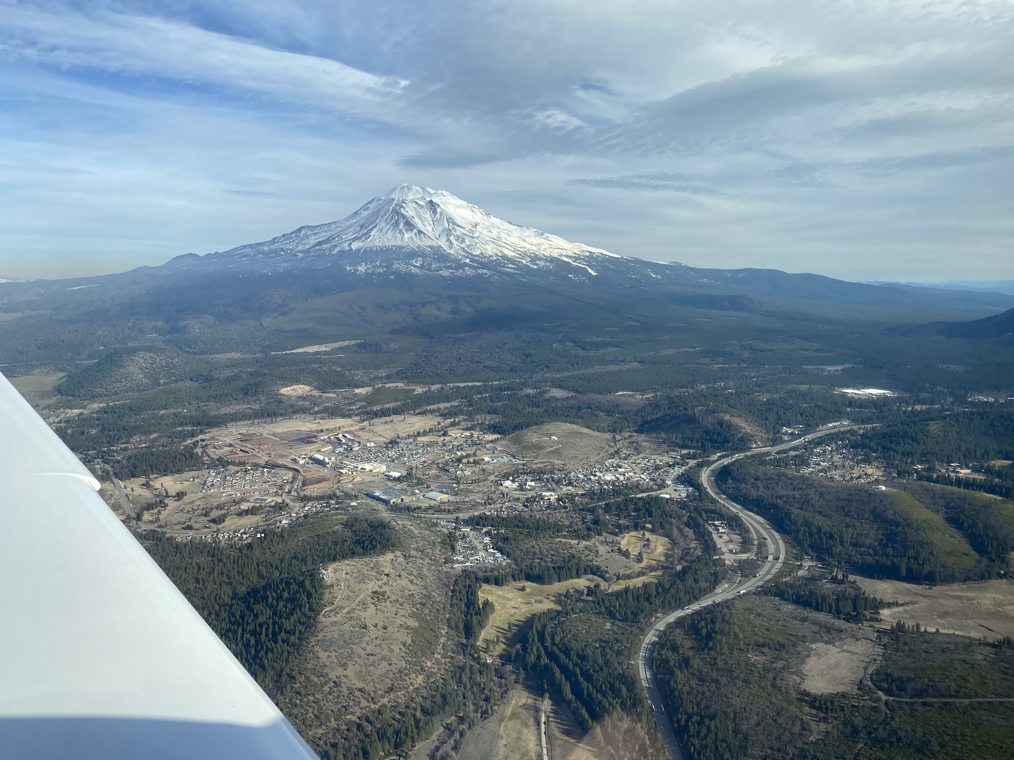 This aerial photograph, taken from an airplane window, shows a snow-capped mountain peak rising above a forested valley with a small town and a winding highway below. The sky is partly cloudy, and the wing of the aircraft is visible in the bottom-left corner.