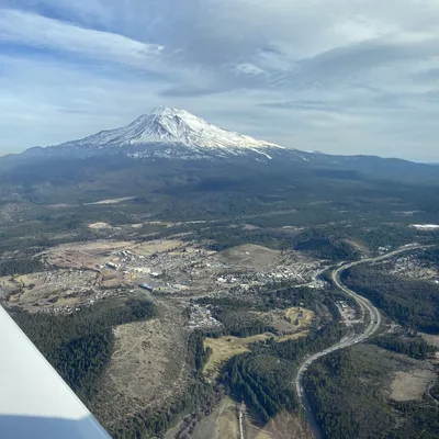 This aerial photograph, taken from an airplane window, shows a snow-capped mountain peak rising above a forested valley with a small town and a winding highway below. The sky is partly cloudy, and the wing of the aircraft is visible in the bottom-left corner.