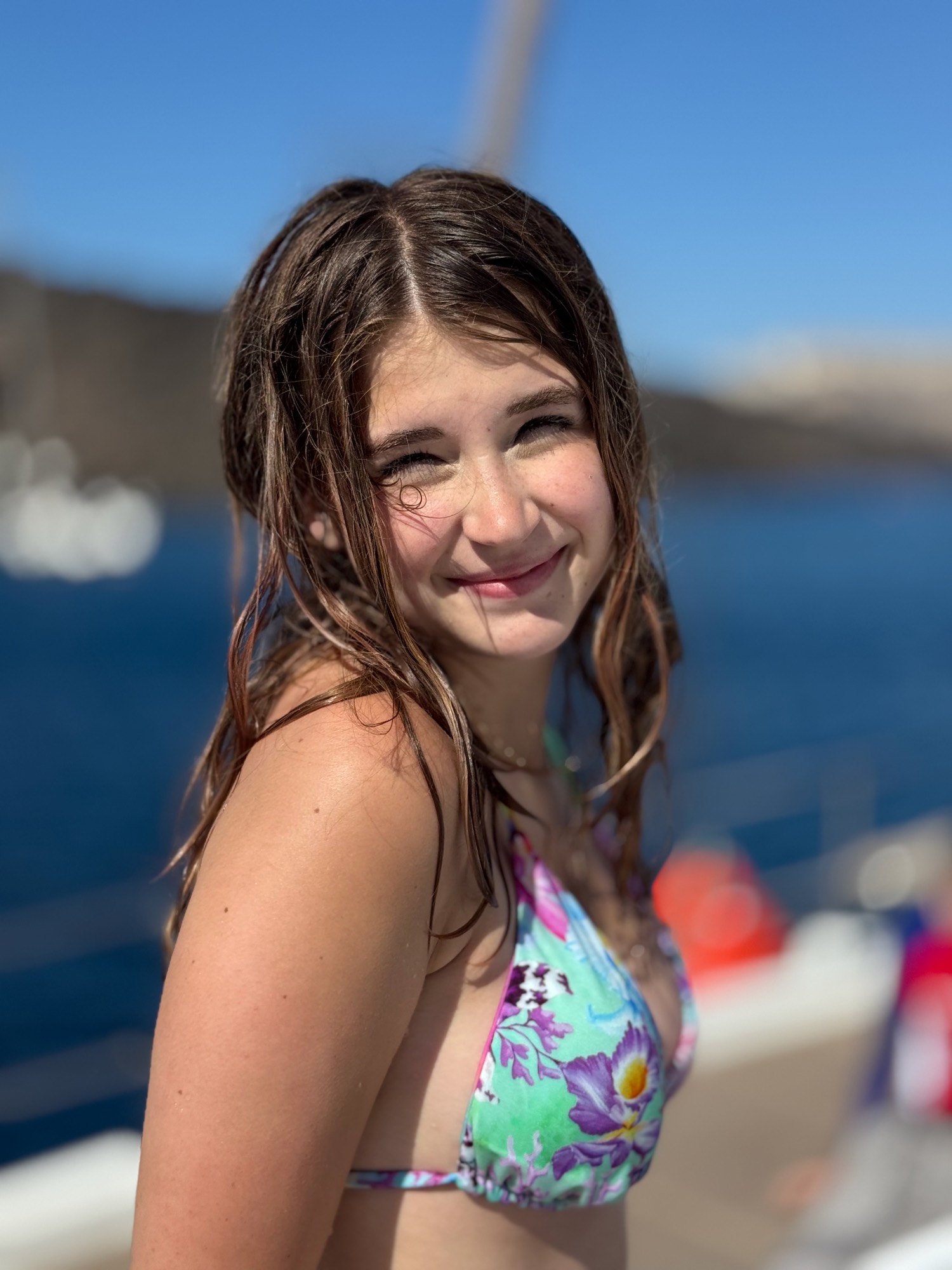 A young woman with wet hair and a floral bikini smiles at the camera while posing outdoors. She is on a boat, with deep blue water and a blurred coastline visible in the background under a clear blue sky.