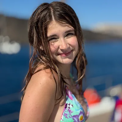 A young woman with wet hair and a floral bikini smiles at the camera while posing outdoors. She is on a boat, with deep blue water and a blurred coastline visible in the background under a clear blue sky.
