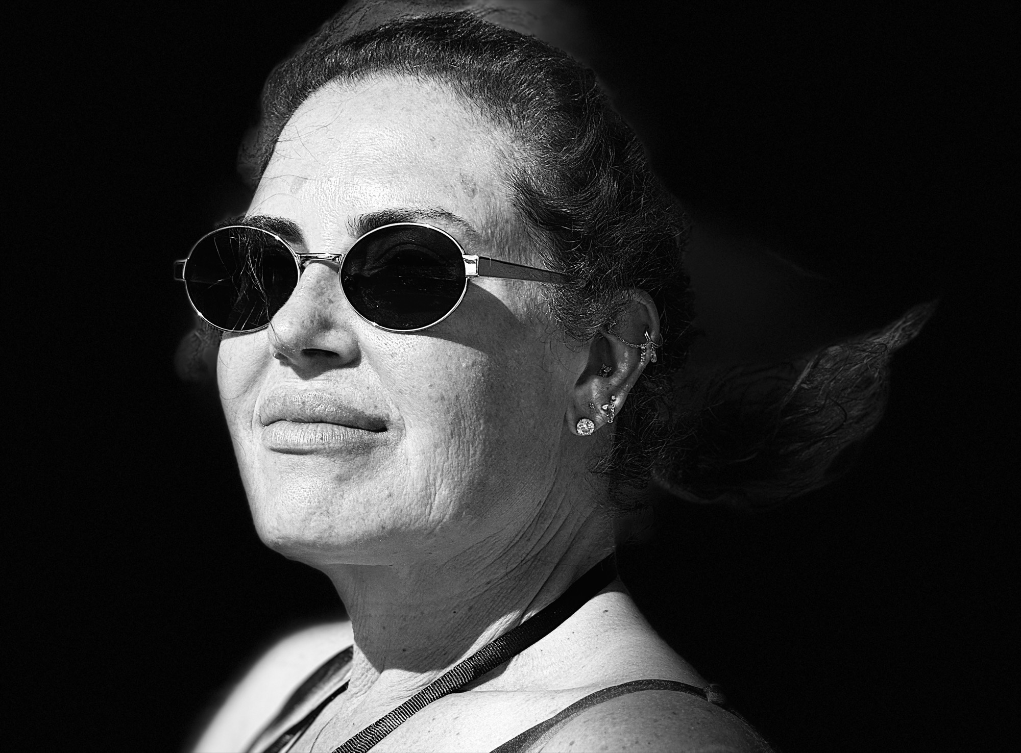 A black and white, close-up portrait shows a woman wearing round sunglasses and multiple ear piercings, looking slightly off-camera with a faint smile. Her hair is pulled back, and she is illuminated by bright sunlight against a dark, shadowed background.