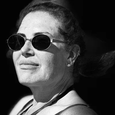 A black and white, close-up portrait shows a woman wearing round sunglasses and multiple ear piercings, looking slightly off-camera with a faint smile. Her hair is pulled back, and she is illuminated by bright sunlight against a dark, shadowed background.