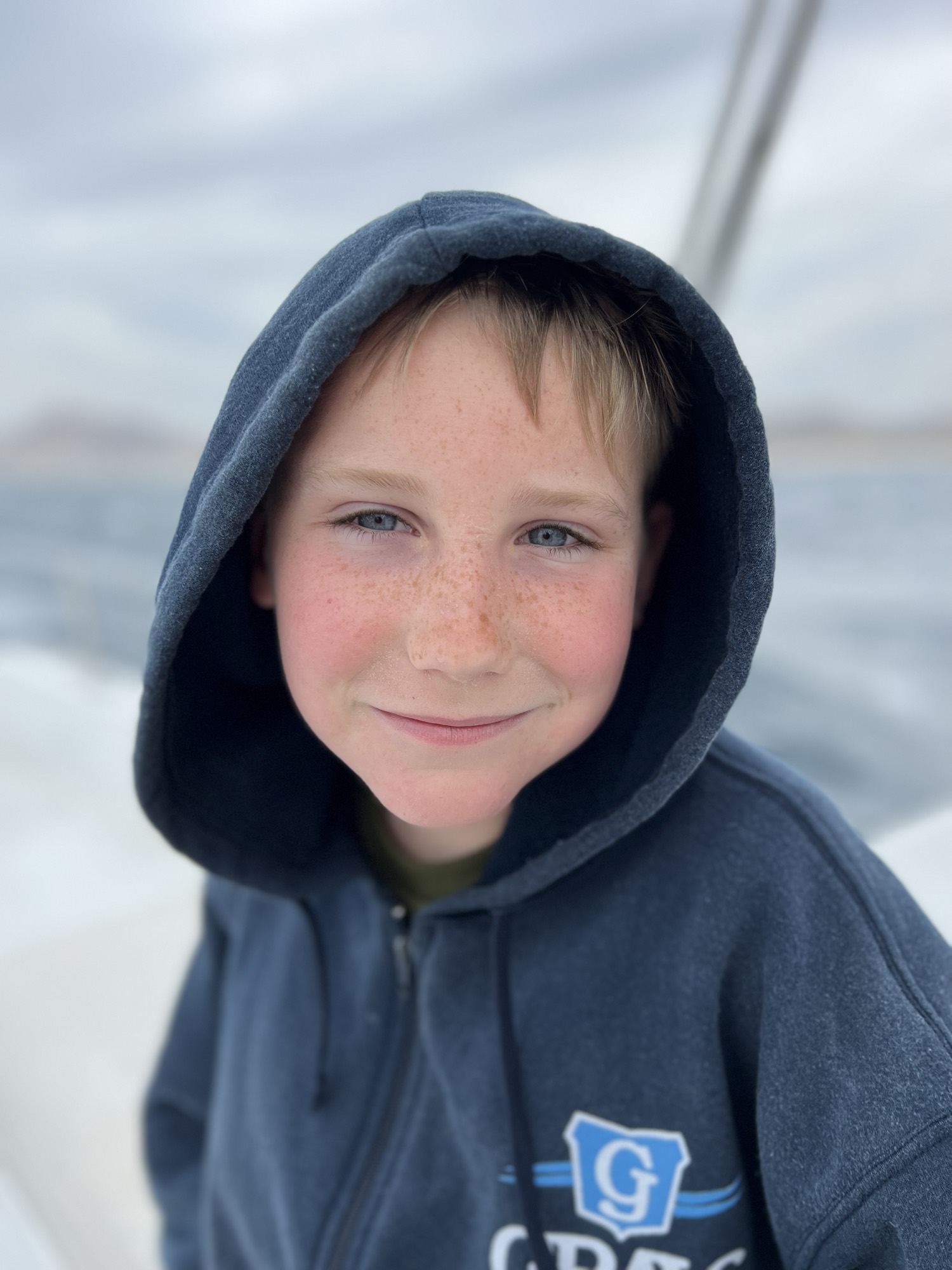 A close-up portrait shows a smiling young boy with freckles wearing a dark blue hooded sweatshirt. He is outdoors on a boat, with the ocean and a cloudy sky visible in the blurred background.