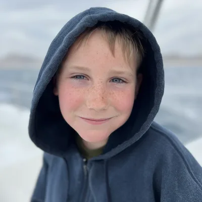 A close-up portrait shows a smiling young boy with freckles wearing a dark blue hooded sweatshirt. He is outdoors on a boat, with the ocean and a cloudy sky visible in the blurred background.