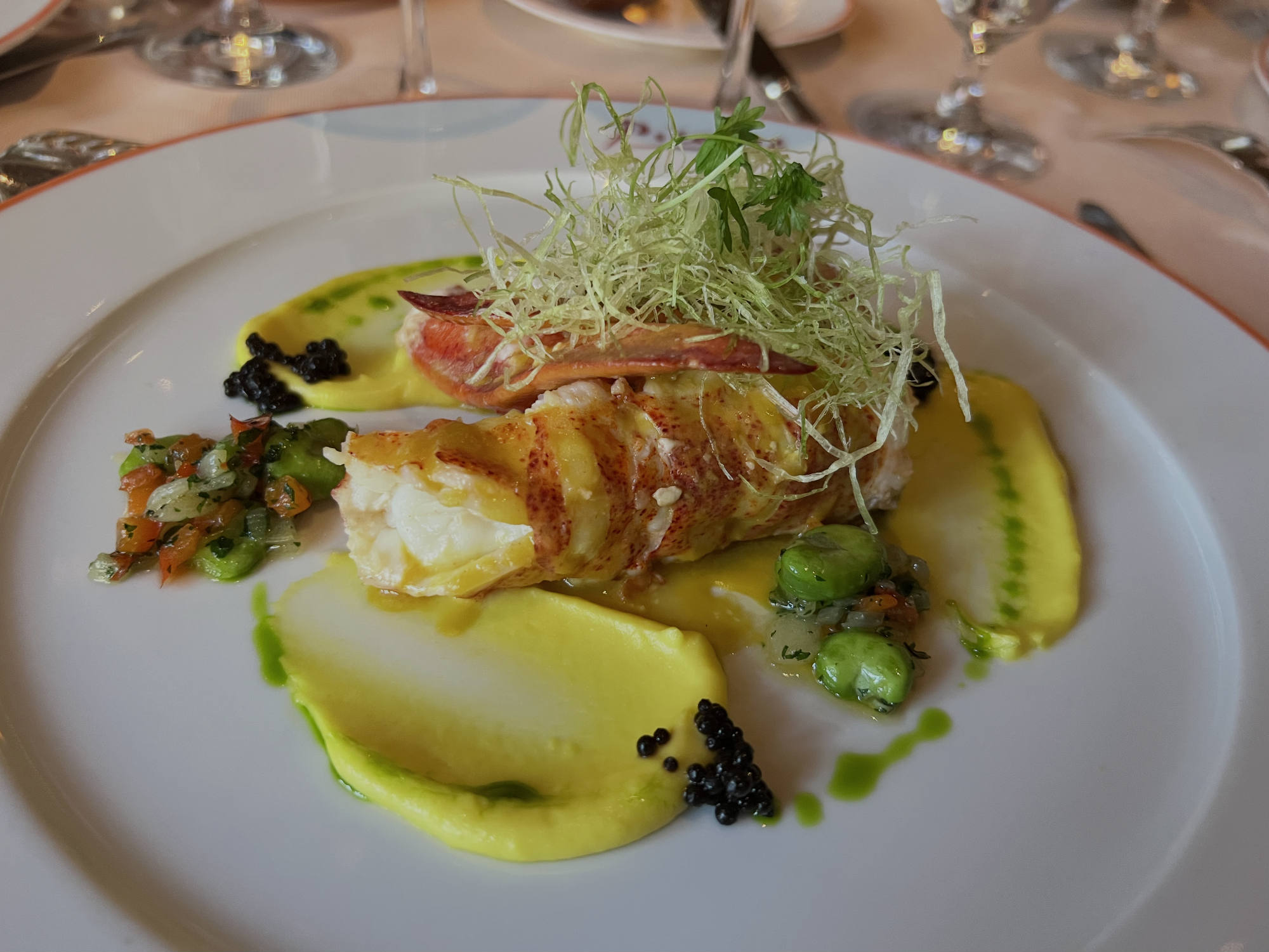 An elegant lobster dish is plated with yellow puree, green herbs, and caviar on a white plate at a restaurant table.