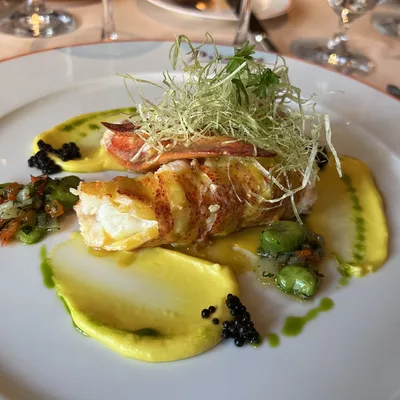 An elegant lobster dish is plated with yellow puree, green herbs, and caviar on a white plate at a restaurant table.
