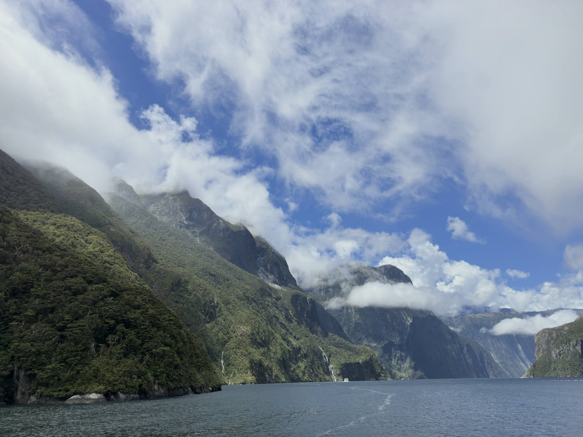 This landscape photograph captures steep, forested mountains rising from a body of water under a blue sky filled with dramatic white clouds. Low-hanging clouds and mist shroud the peaks of the mountains, and a small boat is visible in the distance on the water.