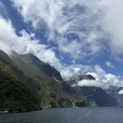 This landscape photograph captures steep, forested mountains rising from a body of water under a blue sky filled with dramatic white clouds. Low-hanging clouds and mist shroud the peaks of the mountains, and a small boat is visible in the distance on the water.