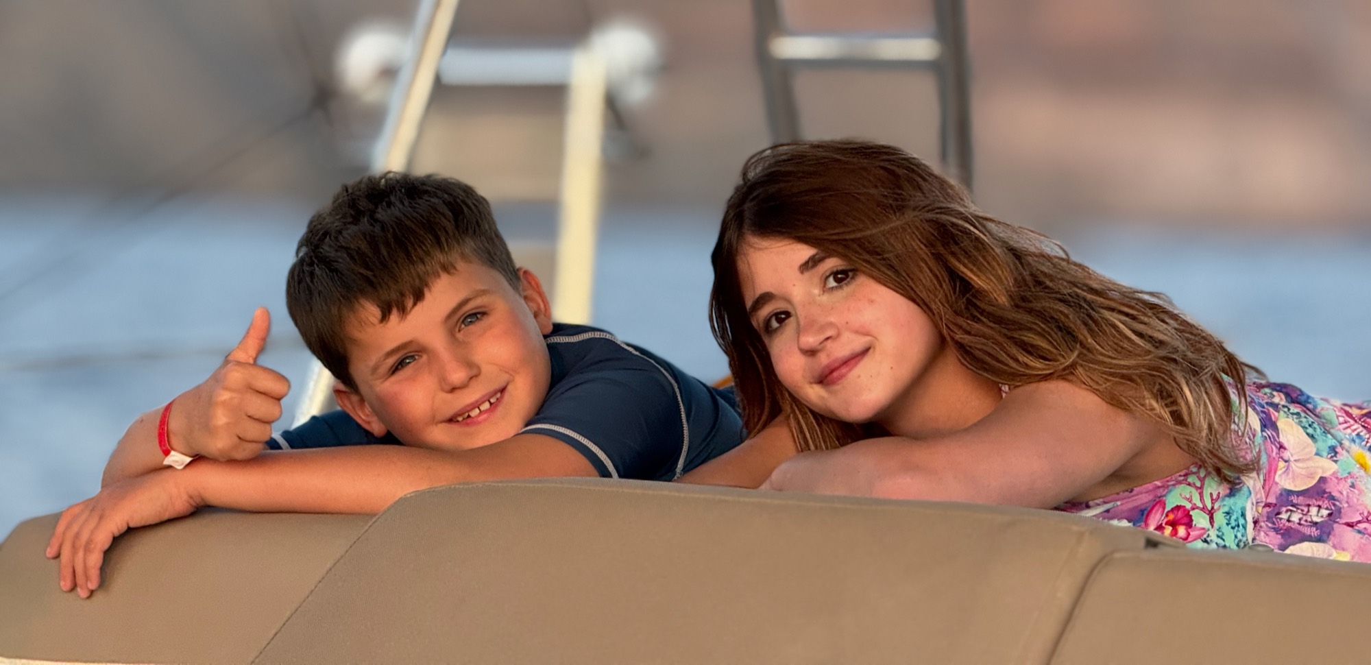 A boy and a girl smile at the camera while leaning on a tan cushion, with the boy giving a thumbs-up. They appear to be outdoors on a boat, with water and metal railings visible in the blurred background.
