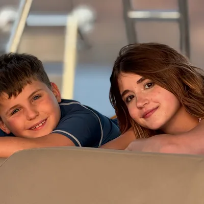 A boy and a girl smile at the camera while leaning on a tan cushion, with the boy giving a thumbs-up. They appear to be outdoors on a boat, with water and metal railings visible in the blurred background.