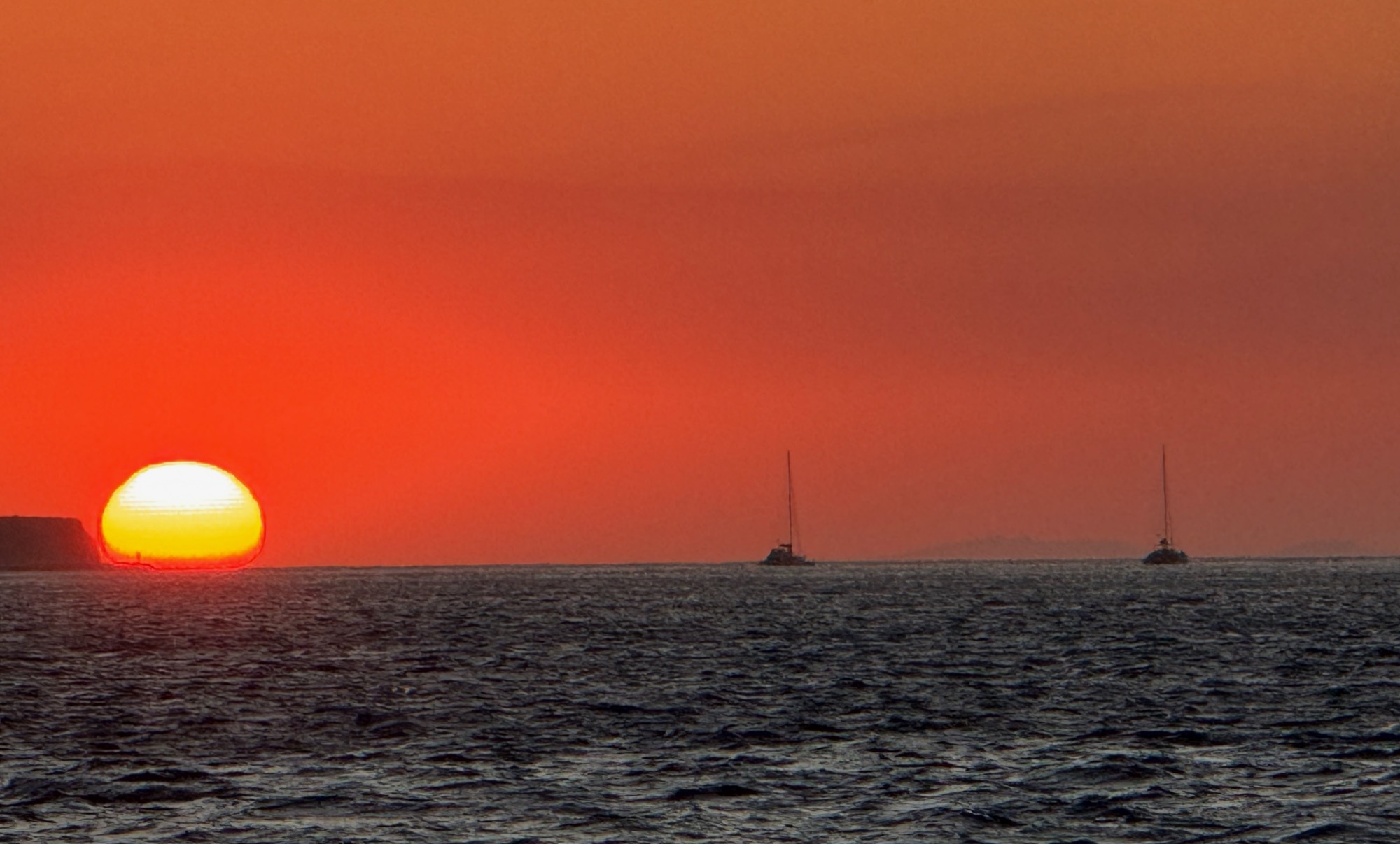 A bright orange sun sets over the ocean horizon next to a distant cliffside, casting a vibrant red glow across the sky. Two sailboats are anchored in the open water under the sunset.