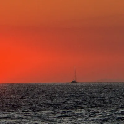 A bright orange sun sets over the ocean horizon next to a distant cliffside, casting a vibrant red glow across the sky. Two sailboats are anchored in the open water under the sunset.