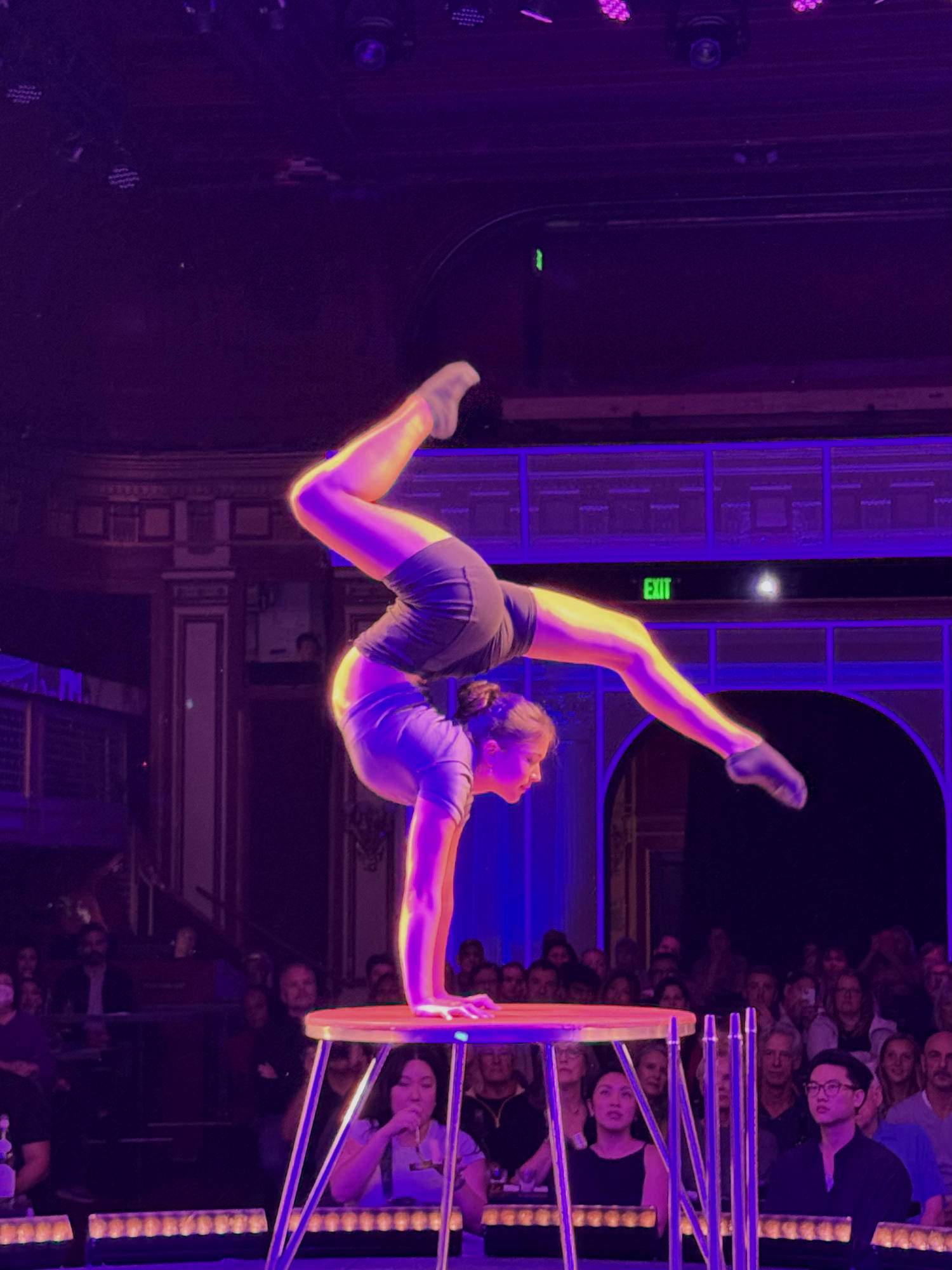 An acrobat performs a handstand contortion on a small, elevated platform in front of a seated audience. The performance takes place on a stage illuminated by purple and pink theatrical lighting.
