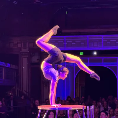 An acrobat performs a handstand contortion on a small, elevated platform in front of a seated audience. The performance takes place on a stage illuminated by purple and pink theatrical lighting.