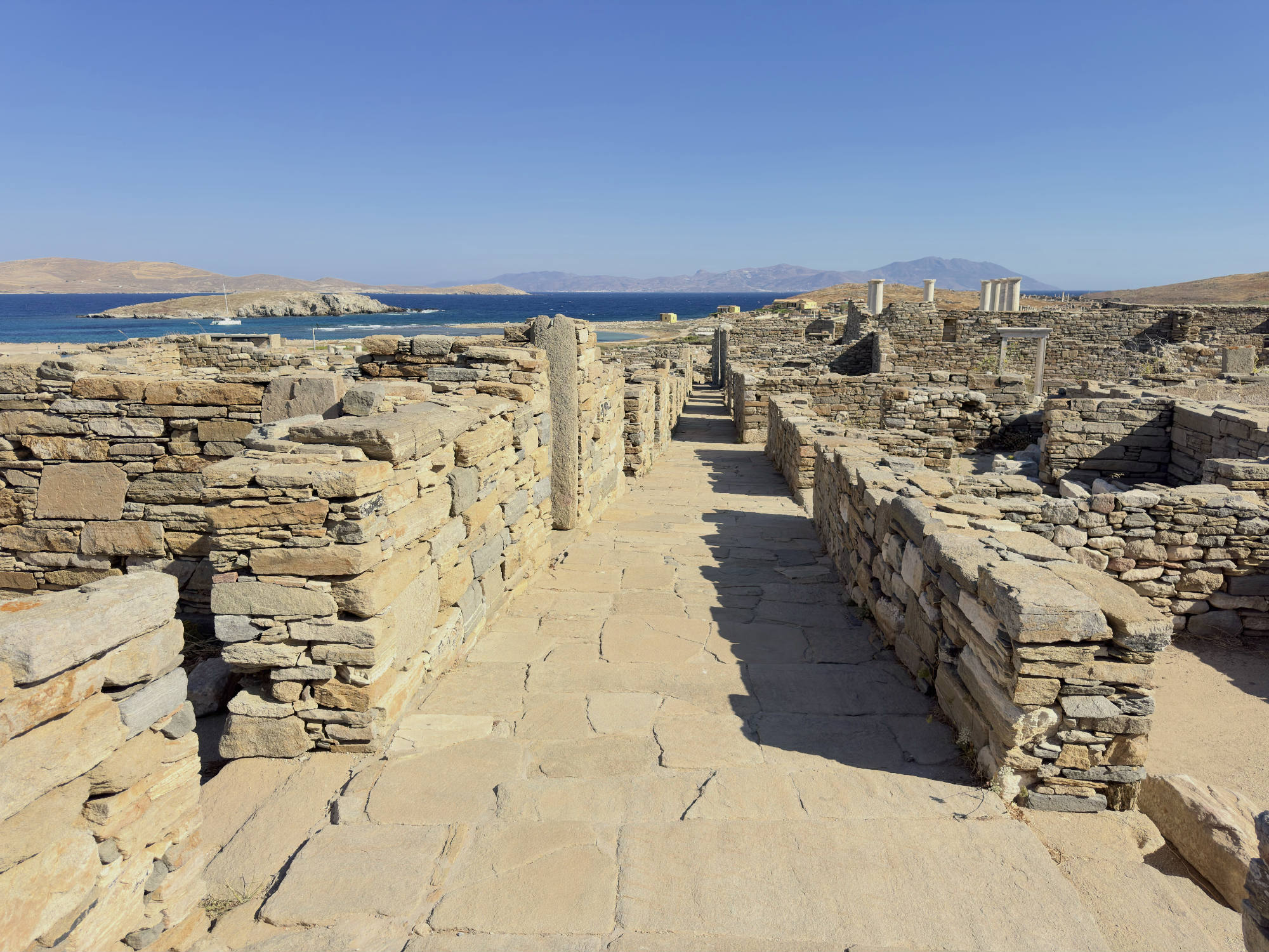 This photo captures a stone-paved pathway leading through ancient stone ruins on a sunny day. In the background, a blue sea with distant islands and mountains is visible under a clear sky.