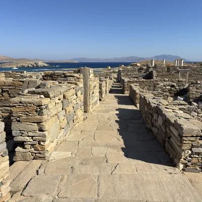 This photo captures a stone-paved pathway leading through ancient stone ruins on a sunny day. In the background, a blue sea with distant islands and mountains is visible under a clear sky.