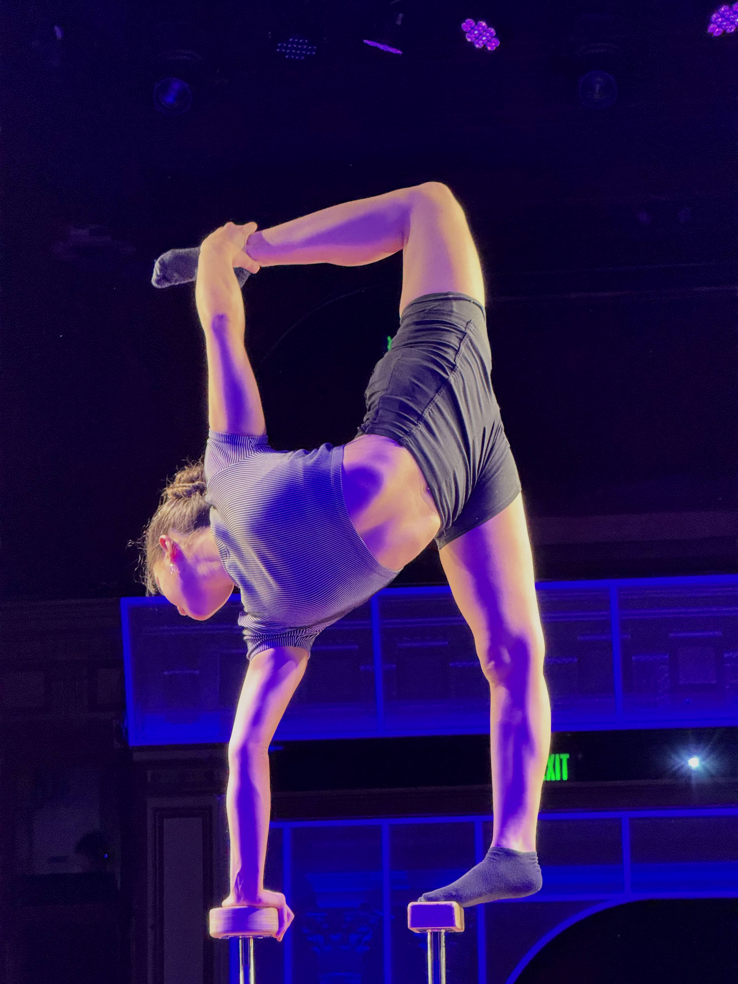 An acrobat performs a flexible handstand-like pose on elevated blocks on a stage. The performance is illuminated by purple and blue stage lighting against a dark background.