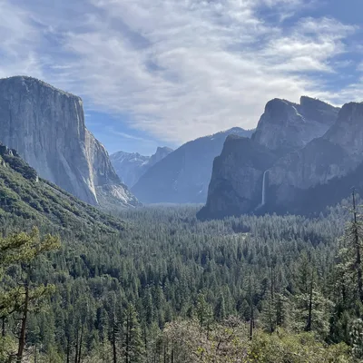 This landscape photograph captures the iconic Tunnel View in Yosemite National Park, featuring El Capitan on the left, Bridalveil Fall on the right, and Half Dome in the distant background. The vast valley is filled with a dense evergreen forest under a partly cloudy blue sky.