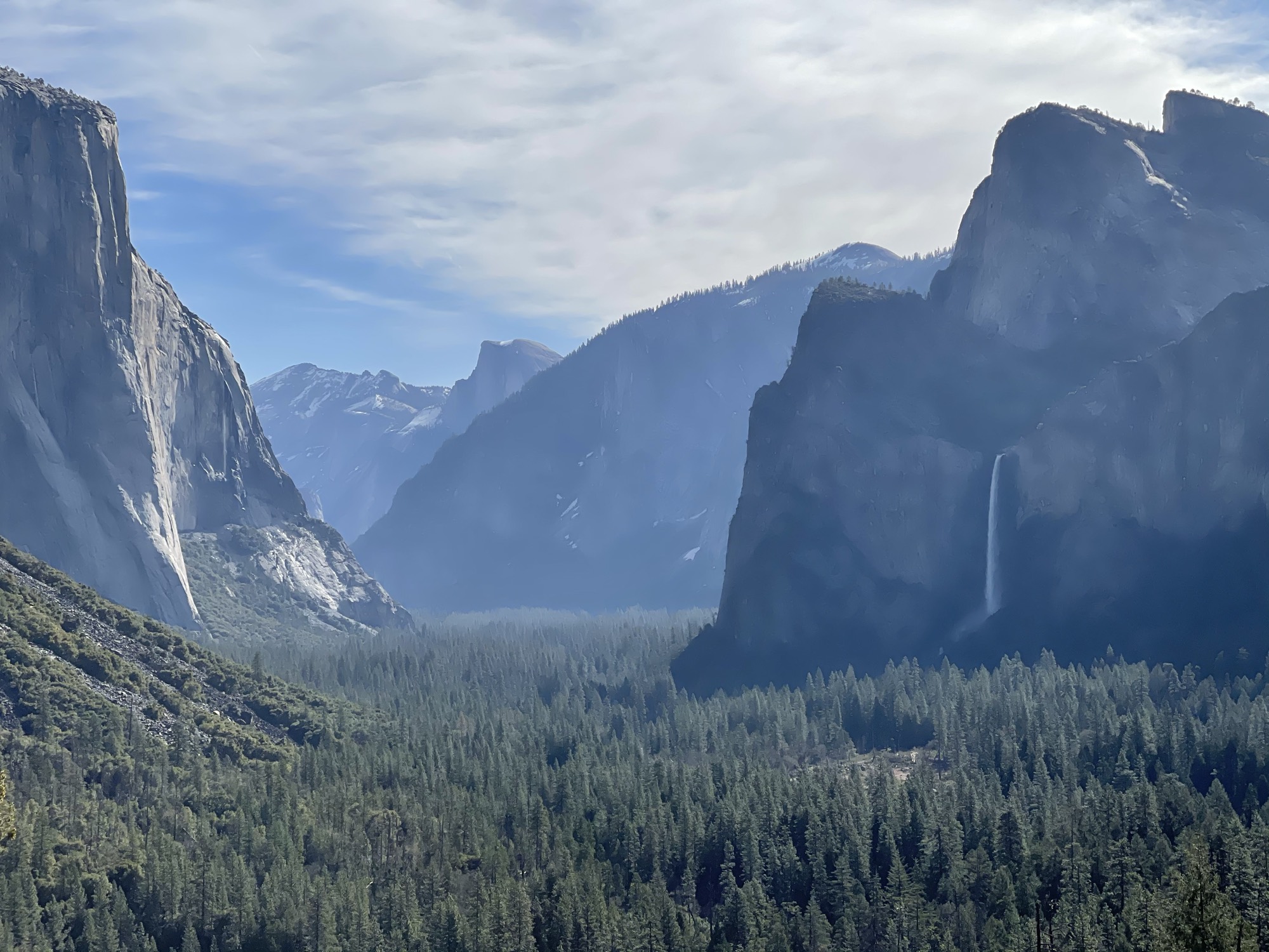 This landscape photograph captures a scenic view of Yosemite Valley, featuring prominent granite cliffs like El Capitan on the left and Bridalveil Fall cascading down a rock face on the right. A dense evergreen forest fills the valley floor below, stretching toward distant, hazy mountain peaks under a partly cloudy sky.