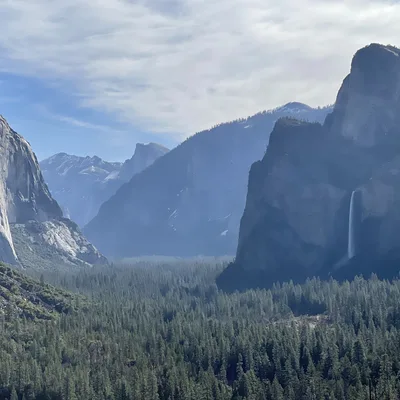 This landscape photograph captures a scenic view of Yosemite Valley, featuring prominent granite cliffs like El Capitan on the left and Bridalveil Fall cascading down a rock face on the right. A dense evergreen forest fills the valley floor below, stretching toward distant, hazy mountain peaks under a partly cloudy sky.