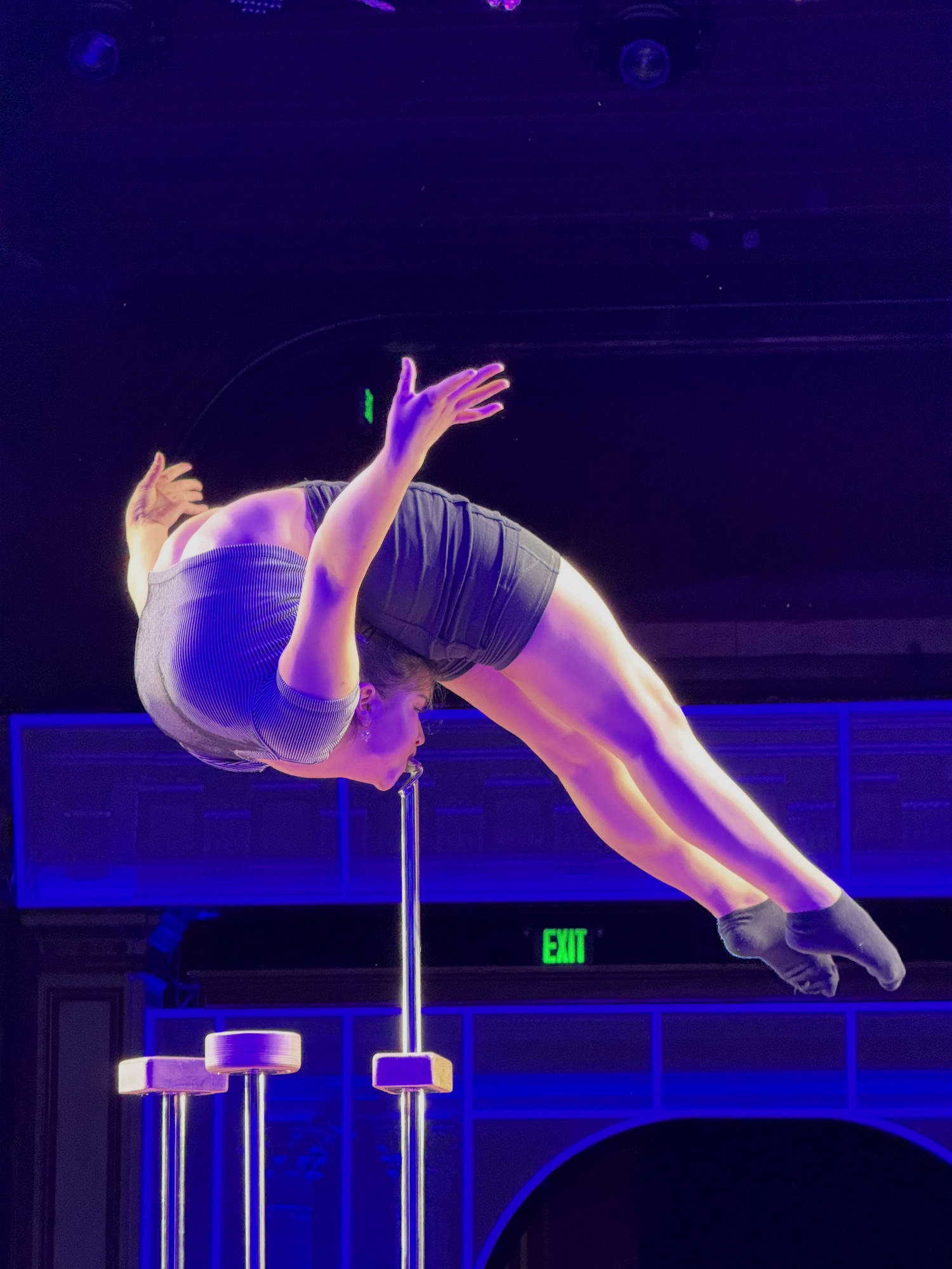 A contortionist performs a mouth hang on a metal stand, bending her body backward on a stage illuminated by blue and purple lights.
