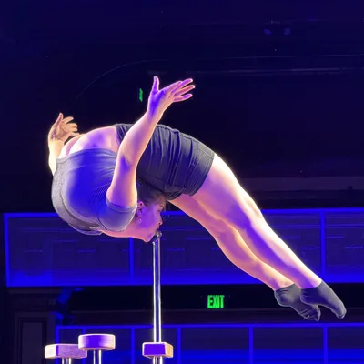 A contortionist performs a mouth hang on a metal stand, bending her body backward on a stage illuminated by blue and purple lights.
