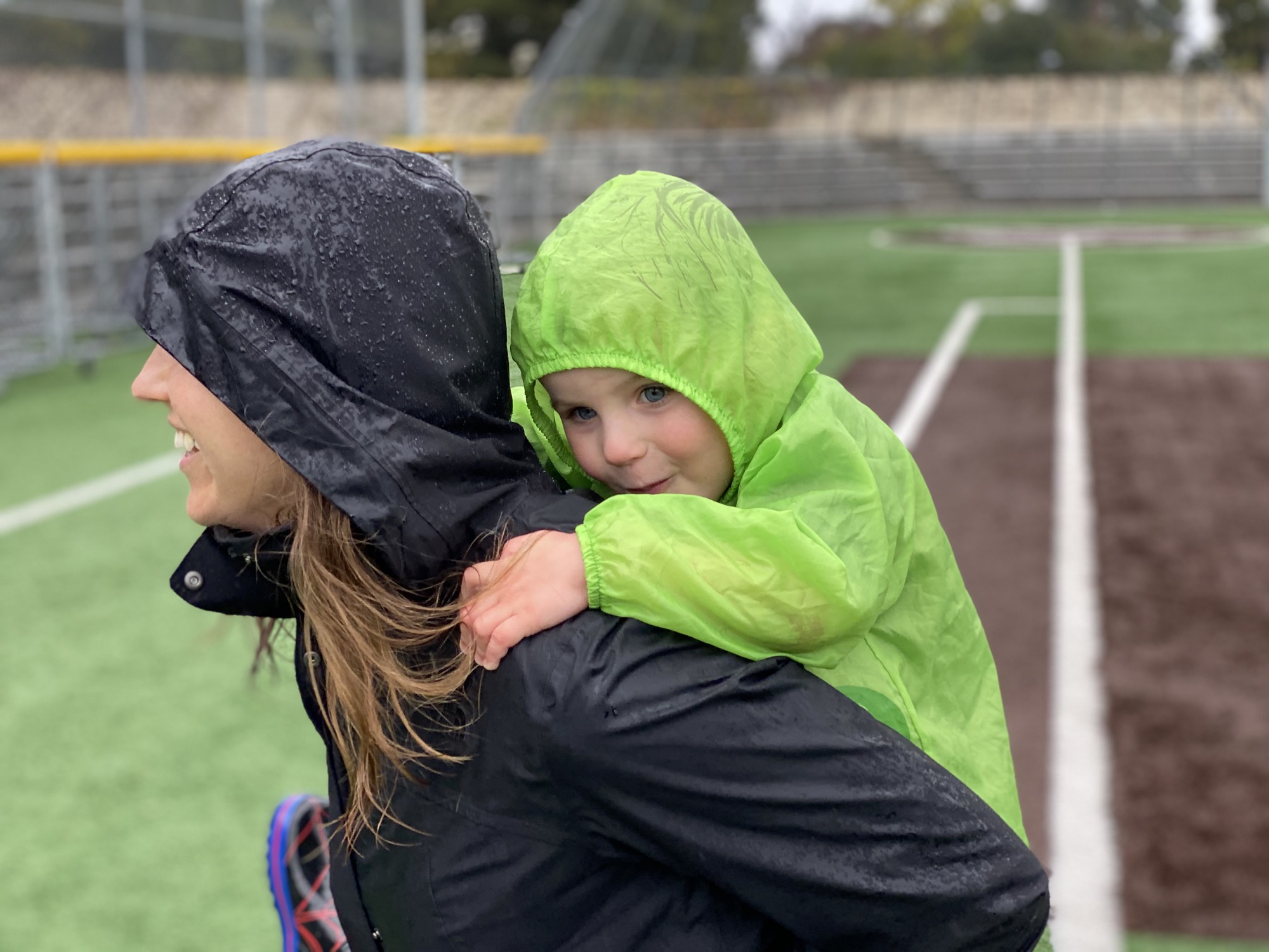 A woman in a wet, black rain jacket gives a young child in a bright green raincoat a piggyback ride on an outdoor sports field. Both individuals have their hoods up in the rainy weather, with bleachers visible in the background.