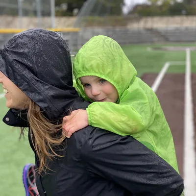 A woman in a wet, black rain jacket gives a young child in a bright green raincoat a piggyback ride on an outdoor sports field. Both individuals have their hoods up in the rainy weather, with bleachers visible in the background.