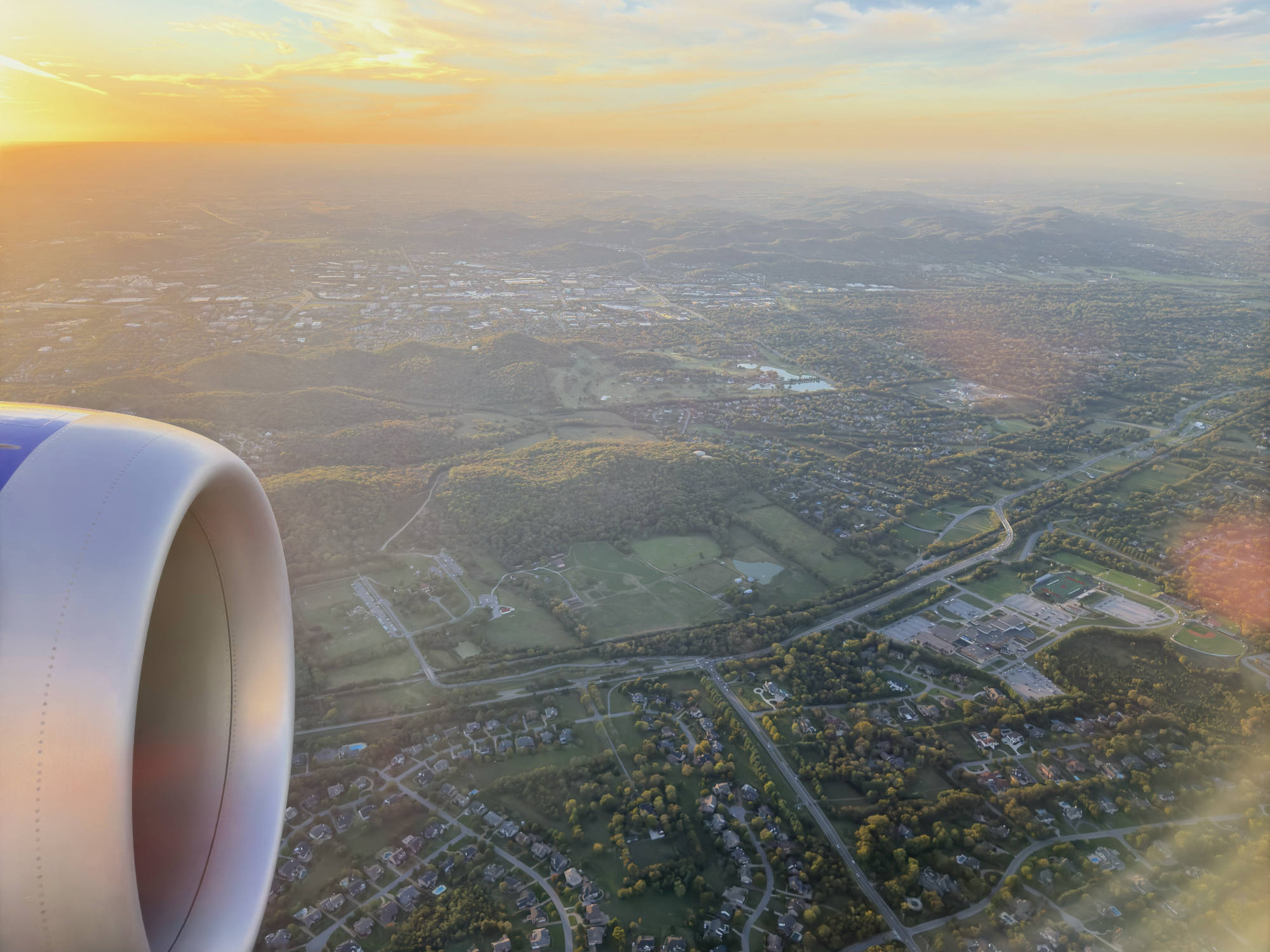 An aerial view taken from an airplane window shows a jet engine in the foreground, looking out over a landscape of suburban neighborhoods, green fields, and rolling hills. The sun is setting on the horizon, casting a warm golden glow across the sky and the land below.
