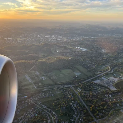 An aerial view taken from an airplane window shows a jet engine in the foreground, looking out over a landscape of suburban neighborhoods, green fields, and rolling hills. The sun is setting on the horizon, casting a warm golden glow across the sky and the land below.
