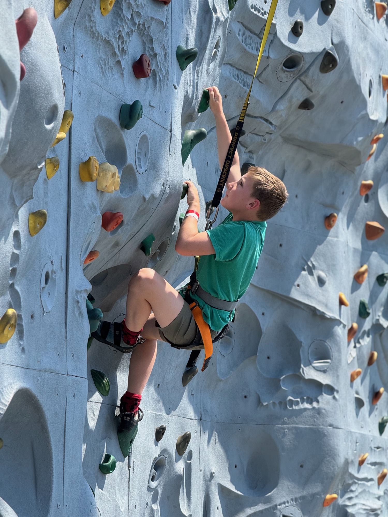 A young boy wearing a green shirt, shorts, and a safety harness climbs an outdoor artificial rock wall. He is holding onto handholds and looking upward as he ascends the textured grey wall.