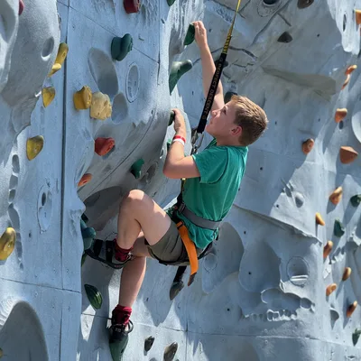 A young boy wearing a green shirt, shorts, and a safety harness climbs an outdoor artificial rock wall. He is holding onto handholds and looking upward as he ascends the textured grey wall.