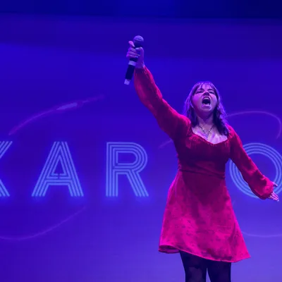 A woman in a red dress sings passionately into a microphone on a stage with purple lighting. Behind her, a large screen displays the word "KARAOKE" in a stylized, glowing font.