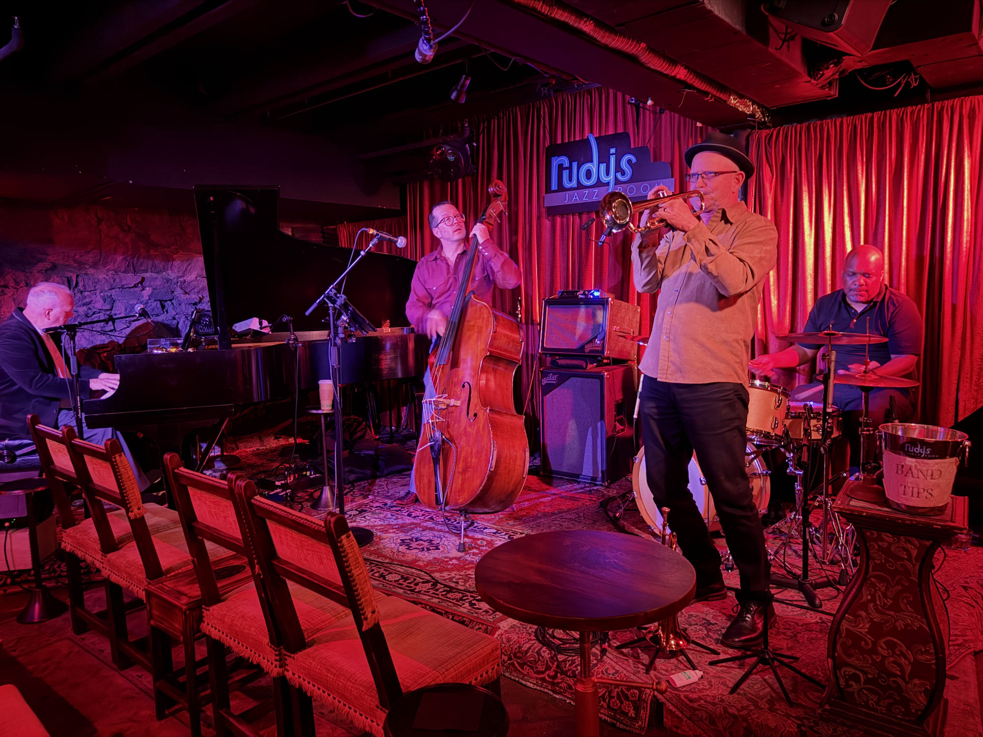 A four-piece jazz band performs on a stage with red curtains and lighting at Rudy's Jazz Room, featuring a pianist, double bassist, trumpeter, and drummer. Empty wooden chairs and a small table are positioned in the foreground of the dimly lit venue.