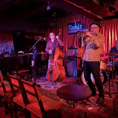 A four-piece jazz band performs on a stage with red curtains and lighting at Rudy's Jazz Room, featuring a pianist, double bassist, trumpeter, and drummer. Empty wooden chairs and a small table are positioned in the foreground of the dimly lit venue.