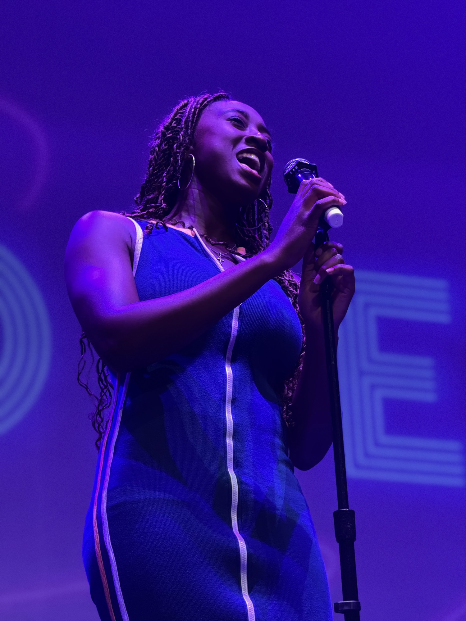 A woman with braided hair wearing a blue dress sings into a microphone on a stage. The scene is illuminated by vibrant purple stage lighting.