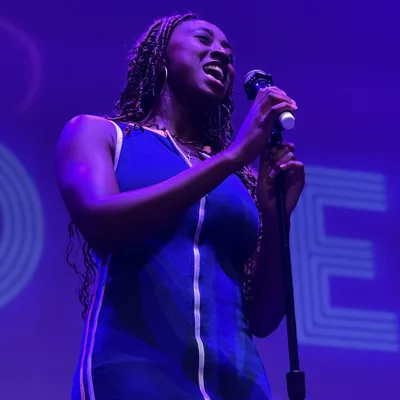 A woman with braided hair wearing a blue dress sings into a microphone on a stage. The scene is illuminated by vibrant purple stage lighting.