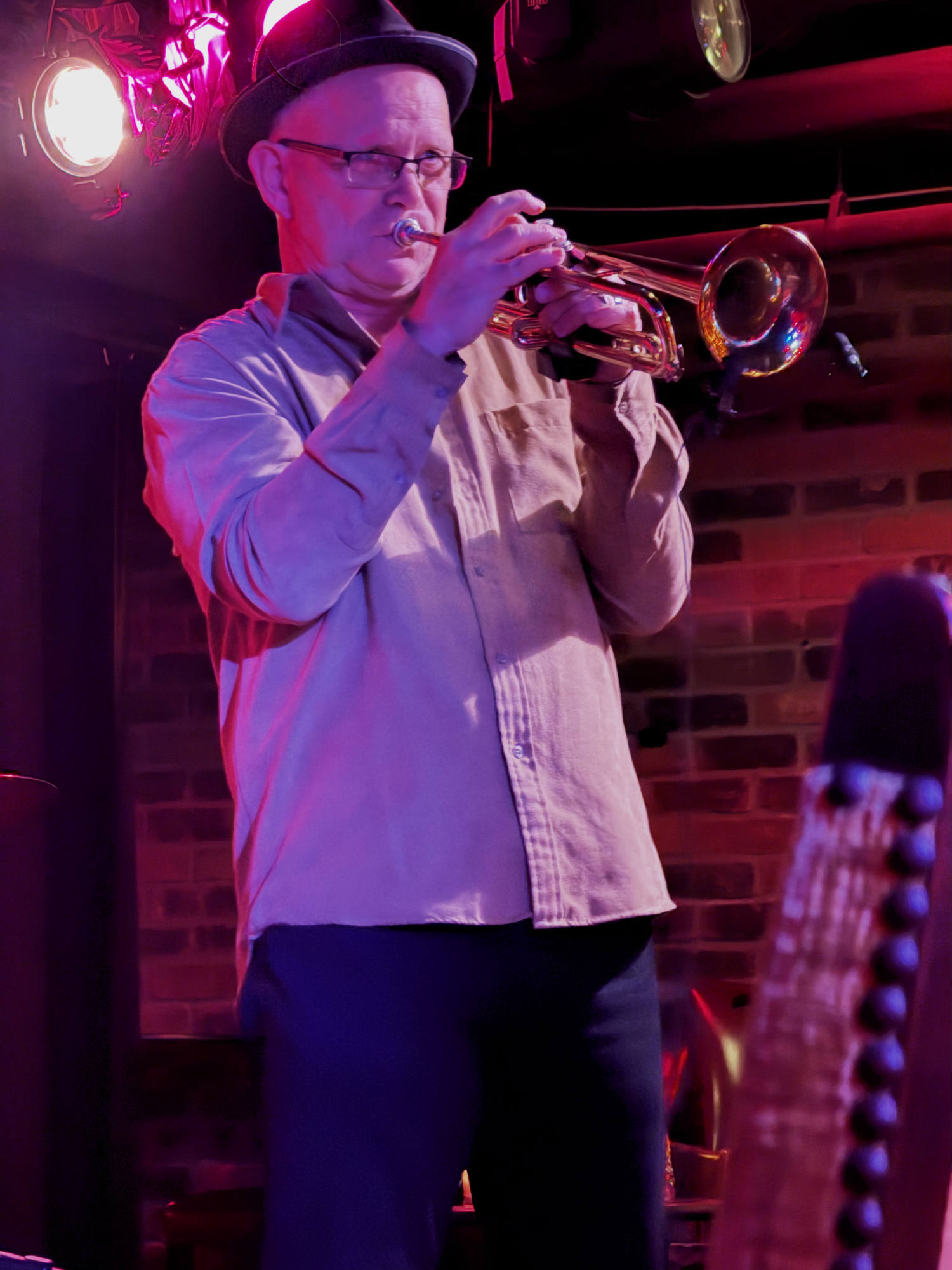 A man wearing a hat and glasses plays a trumpet on a stage with a brick wall background. The scene is illuminated by red stage lighting, with the headstock of a stringed instrument visible in the foreground.