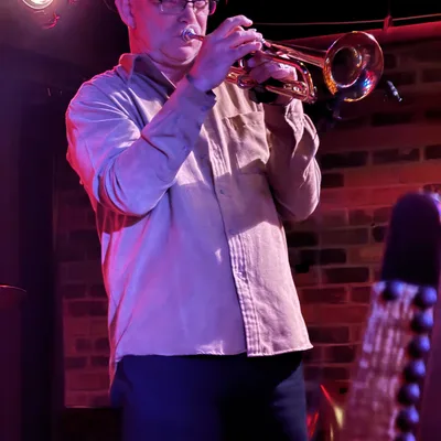 A man wearing a hat and glasses plays a trumpet on a stage with a brick wall background. The scene is illuminated by red stage lighting, with the headstock of a stringed instrument visible in the foreground.