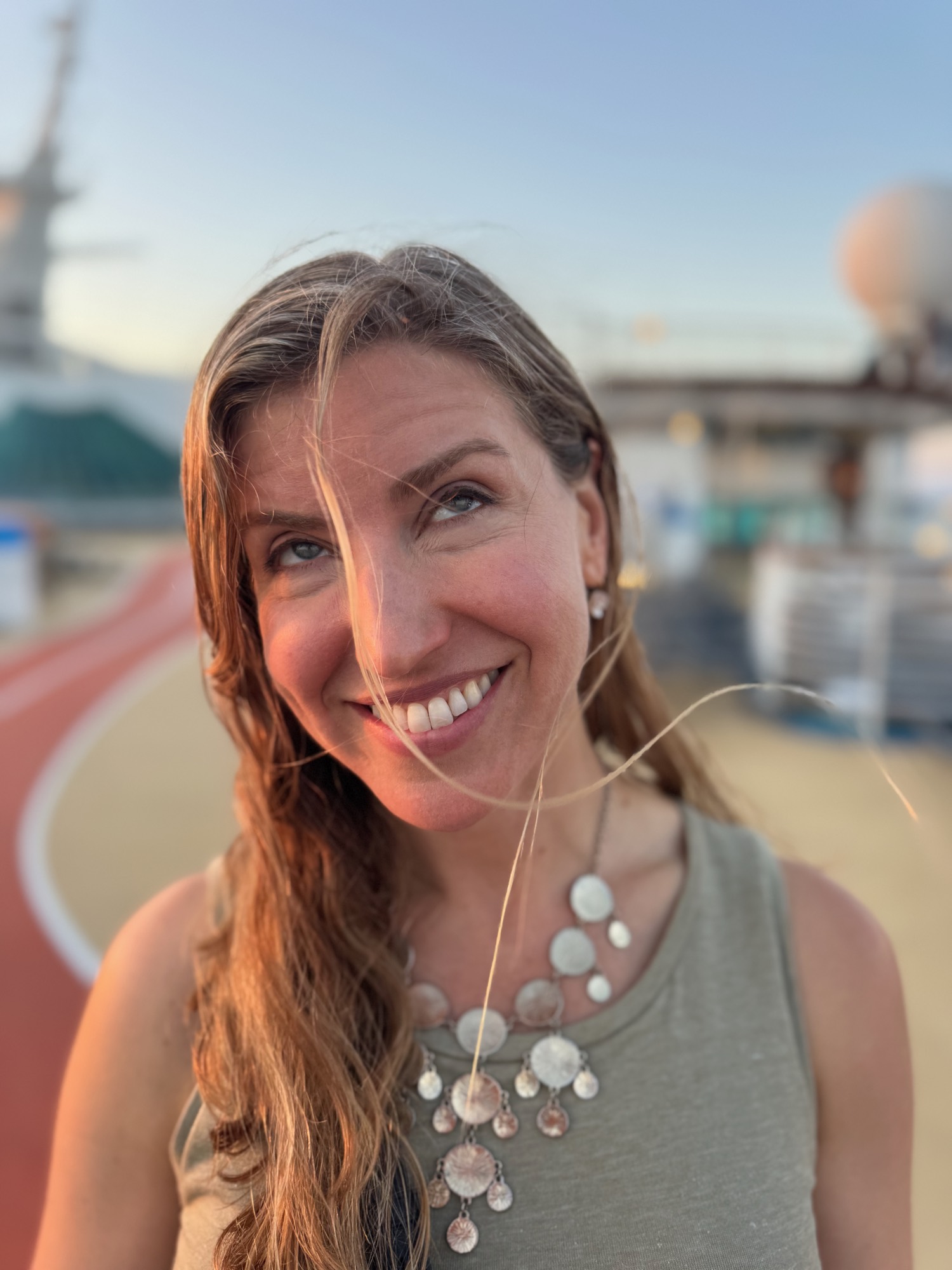 A close-up portrait shows a smiling woman looking upward with her hair blowing in the wind. She is standing outdoors on the deck of a cruise ship, with a running track and ship structures visible in the blurred background.