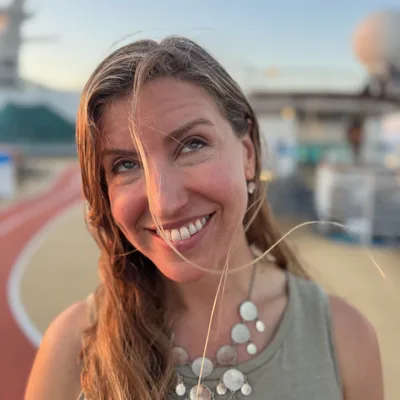 A close-up portrait shows a smiling woman looking upward with her hair blowing in the wind. She is standing outdoors on the deck of a cruise ship, with a running track and ship structures visible in the blurred background.