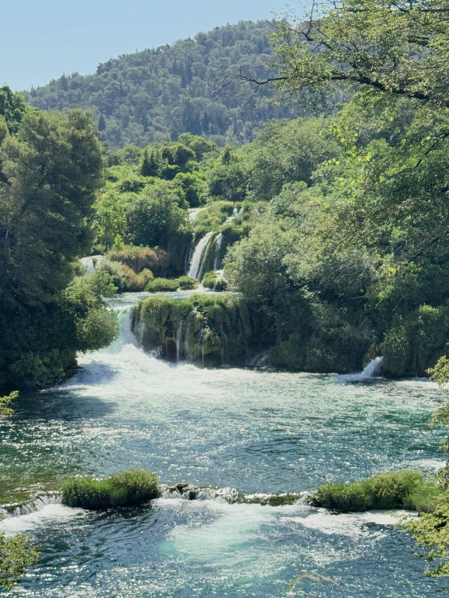 This outdoor landscape photograph features a series of cascading waterfalls flowing into a wide, sunlit river. The water is surrounded by lush green trees and vegetation, with a forested hill rising in the background under a clear blue sky.
