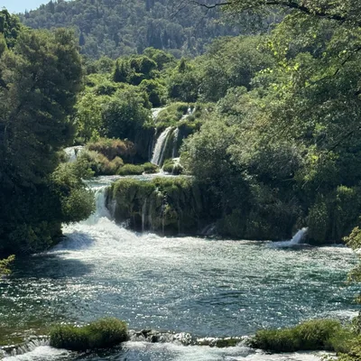 This outdoor landscape photograph features a series of cascading waterfalls flowing into a wide, sunlit river. The water is surrounded by lush green trees and vegetation, with a forested hill rising in the background under a clear blue sky.
