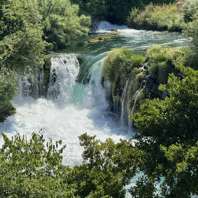 A scenic waterfall cascades over mossy rocks and lush vegetation into a churning pool of turquoise water. The natural landscape is surrounded by dense green trees and foliage under bright daylight.