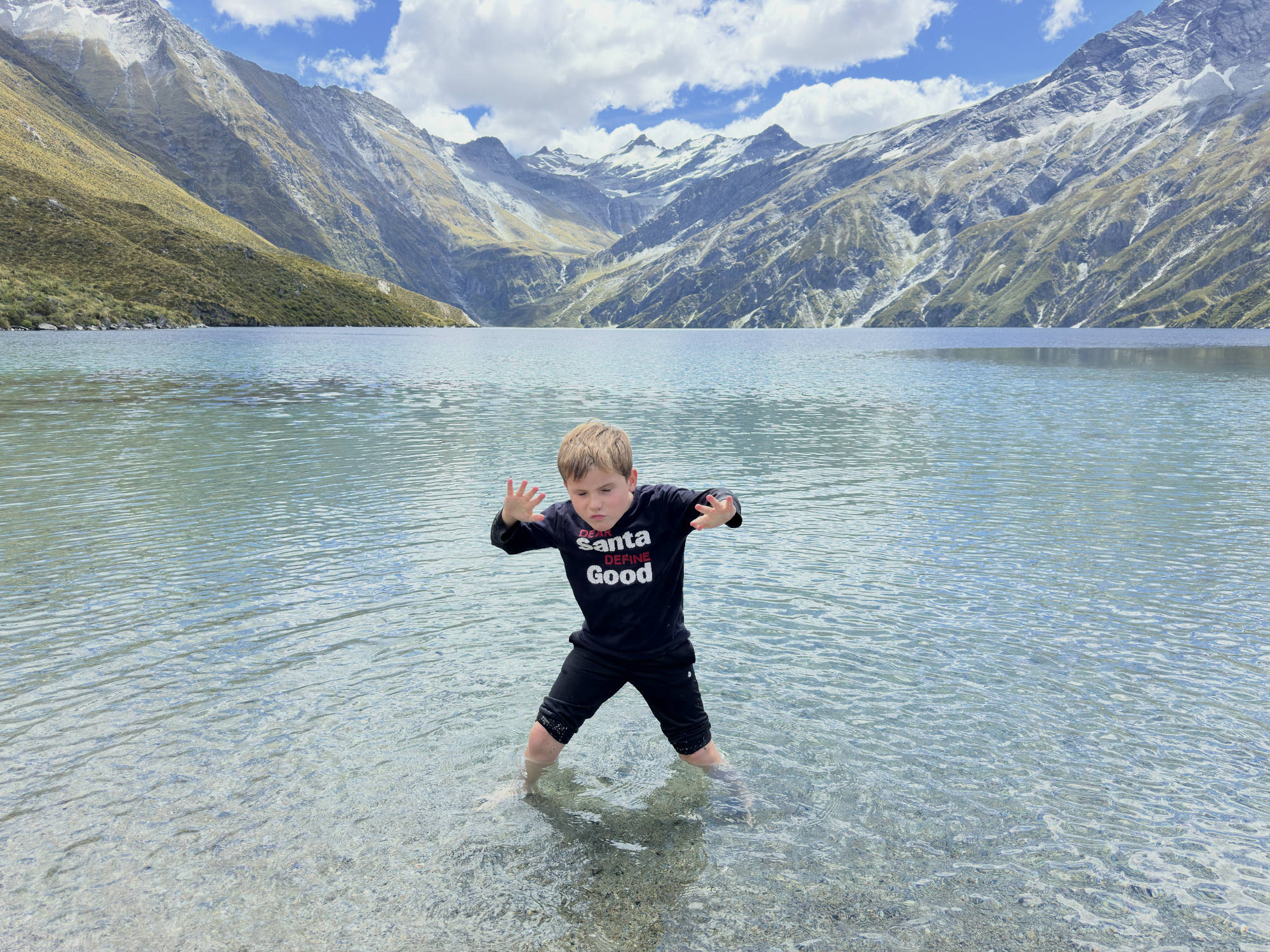 A young boy stands in the shallow, clear water of a mountain lake, posing with his hands raised and a playful expression. In the background, large, snow-dusted mountains rise under a partly cloudy blue sky.